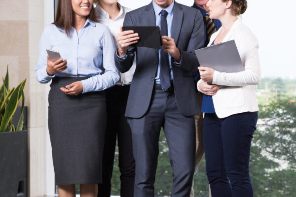 Group of five cheerful business people standing in office lobby, looking at digital tablet computer in his young leaders hands and smiling